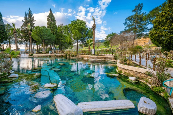 "Cleopatra’s Pool in Pamukkale, Turkey, a natural thermal spring surrounded by trees and ancient Roman ruins with crystal-clear turquoise waters"