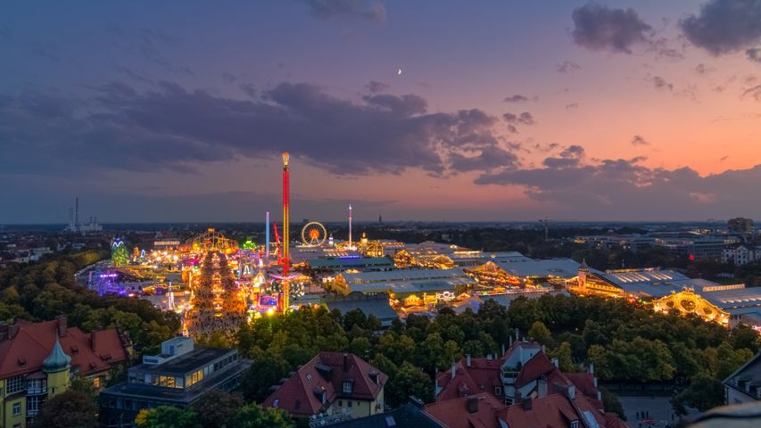 "Aerial view of Oktoberfest fairgrounds in Munich, Germany illuminated at twilight, featuring carnival rides, beer tents, and festive lights under a colorful sunset sky" Oktoberfest in Munich