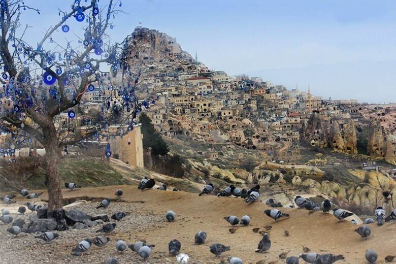 Cappadocia fairy chimneys: "Uçhisar Castle and Pigeon Valley in Cappadocia, Turkey, with pigeons on the ground and a tree decorated with blue evil eye charms overlooking the cave houses"