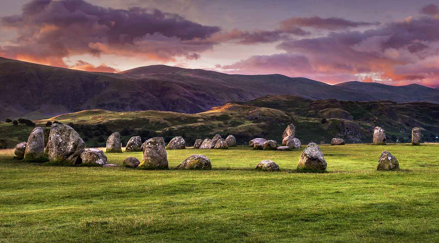 "Castlerigg Stone Circle in the Lake District, England, surrounded by green hills and dramatic clouds at sunset, showcasing ancient stones and scenic countryside"