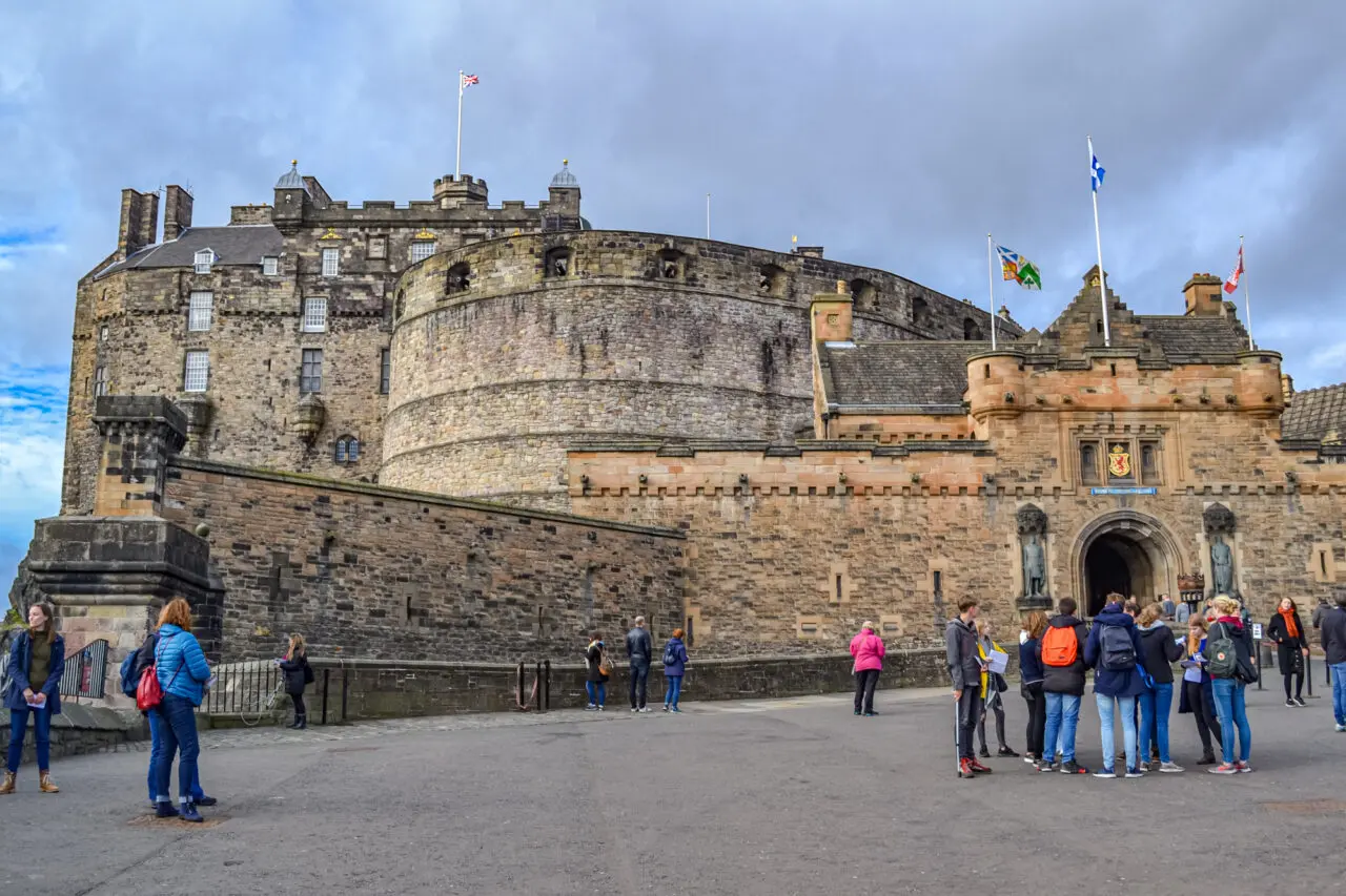 Edinburgh Castle Scotland: "Tourists exploring the entrance of Edinburgh Castle in Scotland with historic stone walls, flying flags, and cloudy blue sky above the fortress."