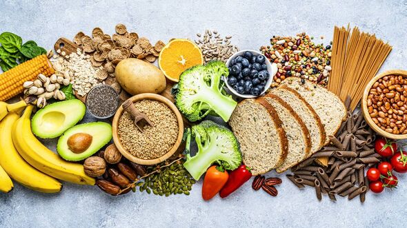 “Assortment of healthy high-fiber foods including whole grains, fruits, vegetables, nuts, seeds, legumes, avocados, bananas, broccoli, and whole-grain bread displayed on a light background.”