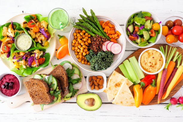 Healthy balanced diet with colorful vegetables, avocado, hummus, whole-grain bread, and smoothies arranged on a white table.